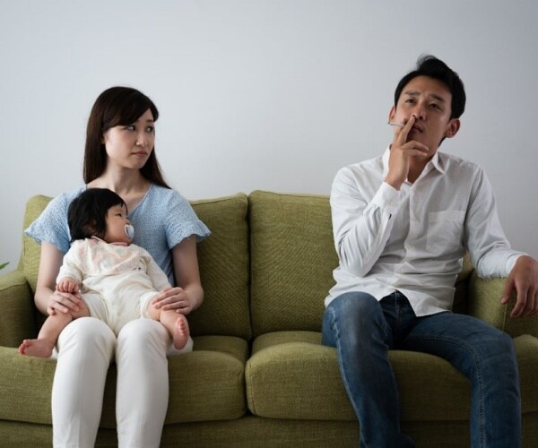 A Mom on couch with baby while the Dad smokes a cigarette at other end of couch