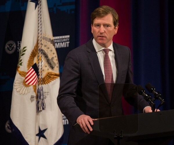 chris krebs speaks in front of a dhs flag, standing at a podium