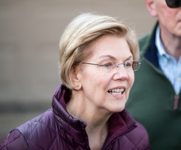 elizabeth warren in a purple jacket speaking to the media with her husband in the background