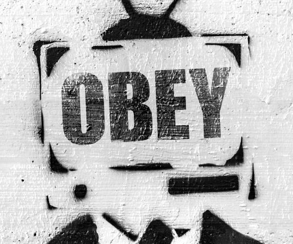 stencil painting on a wall of a man in a suit with a television for a head with the word obey on the screen