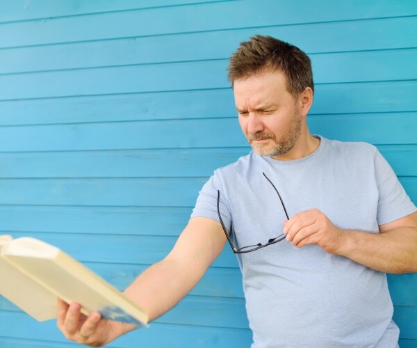 man with glasses in his hands squinting while trying to read a book he is holding far away