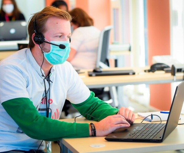 man wearing a face mask and headset looks on a laptop