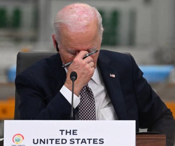 joe biden sitting at a table with his hand over this face and a card reading united states of america on the table