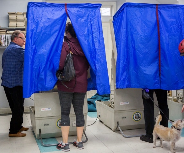 man standing inside a voting booth with blue plastic around him holds a dog and woman votes next to him